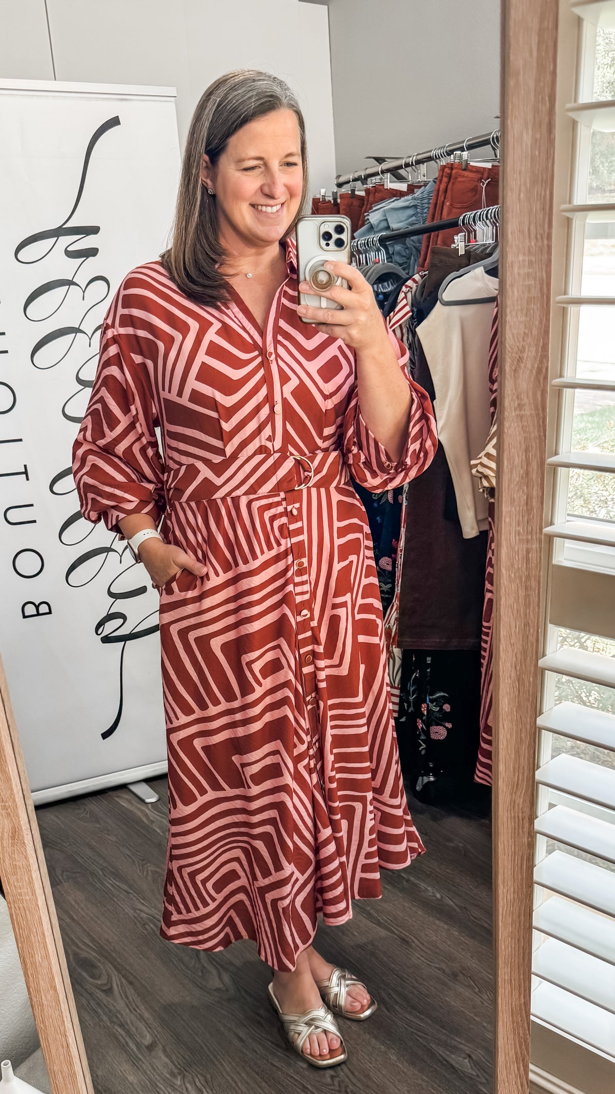 A woman takes a mirror selfie in a boutique showing a rust red and white geometric patterned midi dress with three-quarter sleeves.