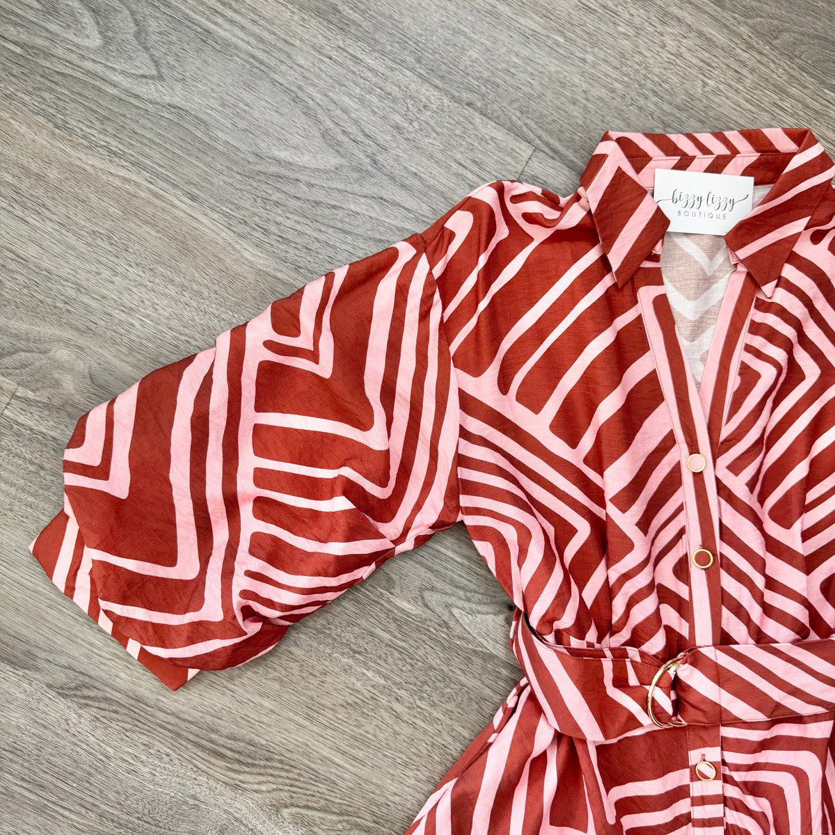 A brown and pink geometric patterned shirt dress with a tie waist and button front, laid flat on a gray wood floor.