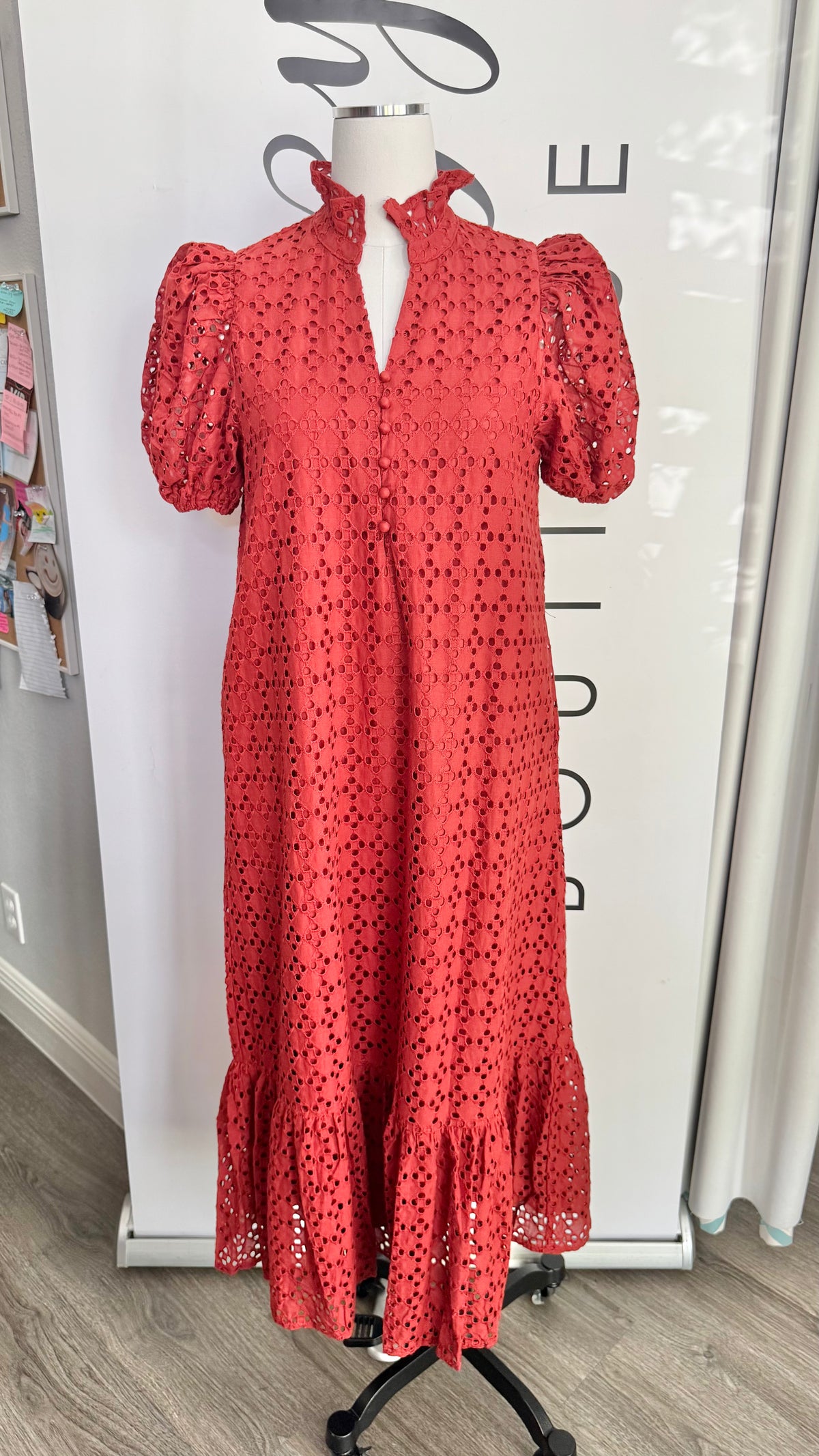 A rust orange eyelet midi dress with puff sleeves and a ruffled hem, displayed on a mannequin in front of a white boutique backdrop.