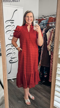 A woman takes a mirror selfie in a boutique wearing a rust orange eyelet cotton  midi dress with puff sleeves and a ruffled hem. The boutique interior is visible behind her, including a clothing rack and a white display board.