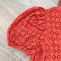 A close-up of a rust orange eyelet fabric with puff sleeves, laid flat on a gray wood-textured surface.