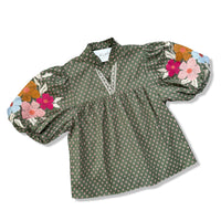 An olive green long-sleeve blouse with a small white geometric pattern, featuring a V-neckline with lace trim and colorful floral embroidery on the puffed sleeves.
