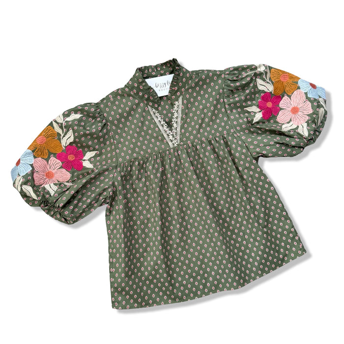 An olive green long-sleeve blouse with a small white geometric pattern, featuring a V-neckline with lace trim and colorful floral embroidery on the puffed sleeves.