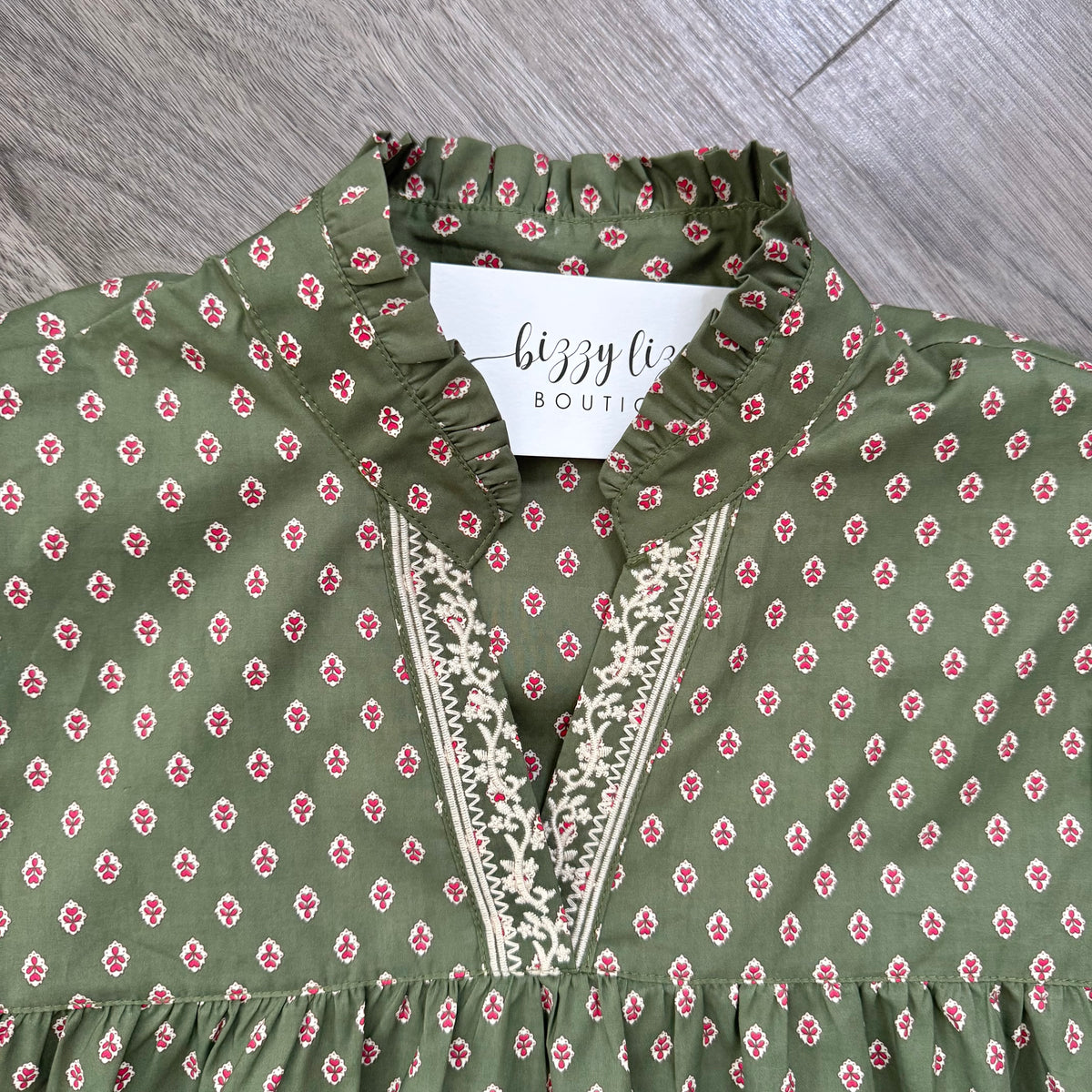 An olive green blouse with a small pink and white floral pattern, featuring a ruffled high collar and a V-neckline with cream embroidered detailing. A white brand tag reading 'bizzy lizzy BOUTIQUE' is visible inside the collar, and the garment is laid flat on a gray wood-textured surface.