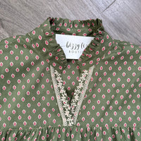 An olive green blouse with a small pink and white floral pattern, featuring a ruffled high collar and a V-neckline with cream embroidered detailing. A white brand tag reading 'bizzy lizzy BOUTIQUE' is visible inside the collar, and the garment is laid flat on a gray wood-textured surface.