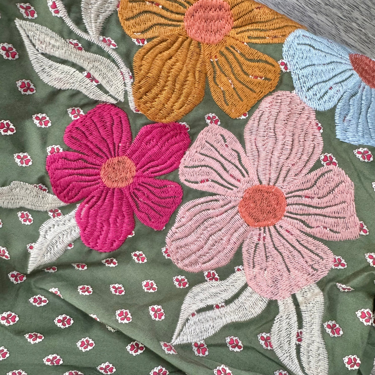 Close-up of embroidered fabric with large floral motifs in pink, orange, and blue on a muted green background, plus small repeating floral prints.