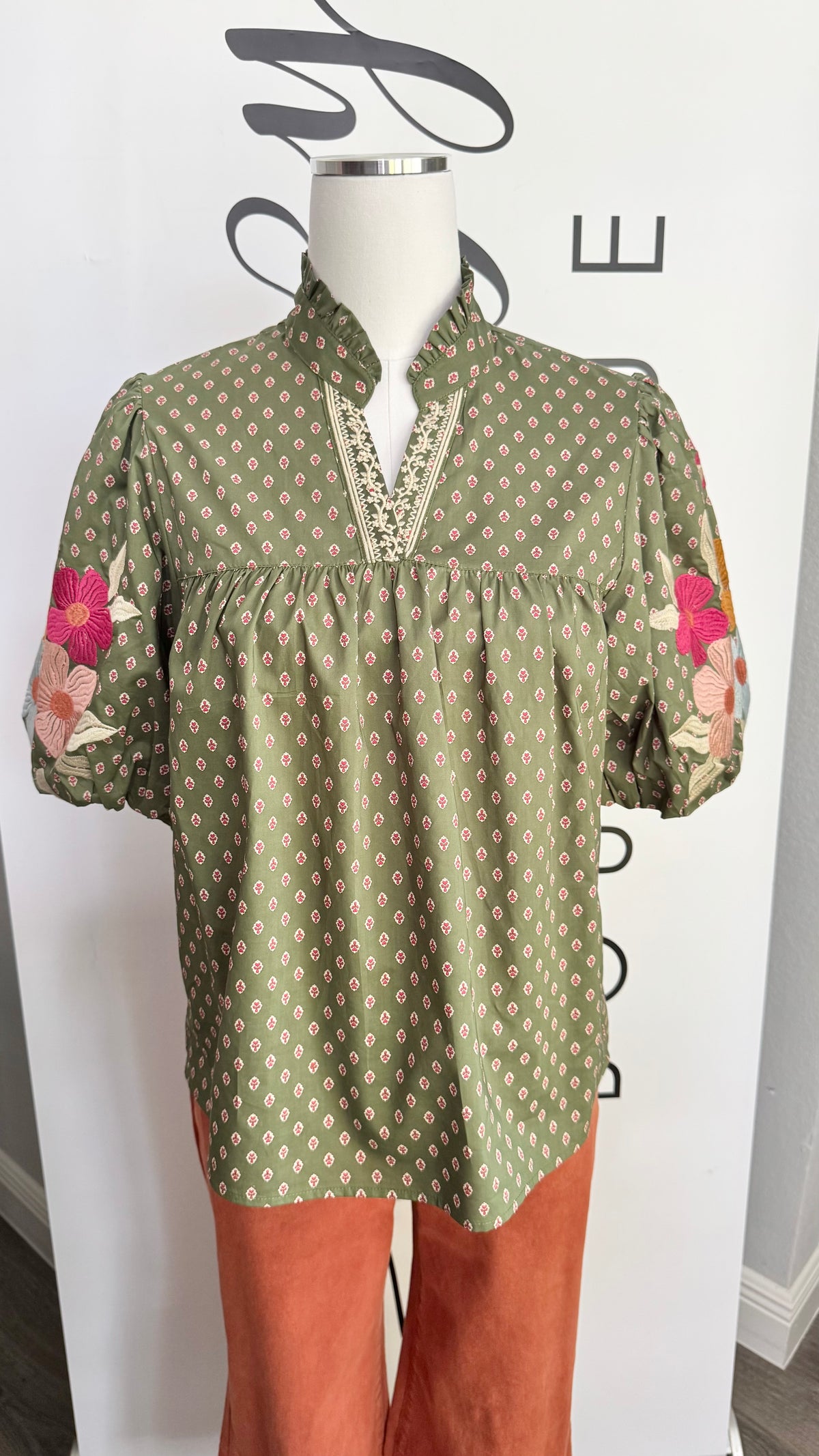 An olive green short-sleeve blouse with a beige geometric pattern and pink floral embroidery on the sleeves, displayed on a mannequin with rust-colored pants against a white branded backdrop.