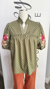 An olive green short-sleeve blouse with a beige geometric pattern and pink floral embroidery on the sleeves, displayed on a mannequin with rust-colored pants against a white branded backdrop.