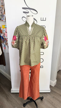A two-piece women's outfit displayed on a mannequin, featuring a sage green short-sleeve top with beige polka dots and pink floral embroidery on the sleeves, paired with rust-orange wide-leg pants.