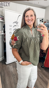 A woman takes a mirror selfie in a boutique wearing an olive green short-sleeve blouse with a white geometric pattern, white floral embroidery on the sleeve, and white pants.