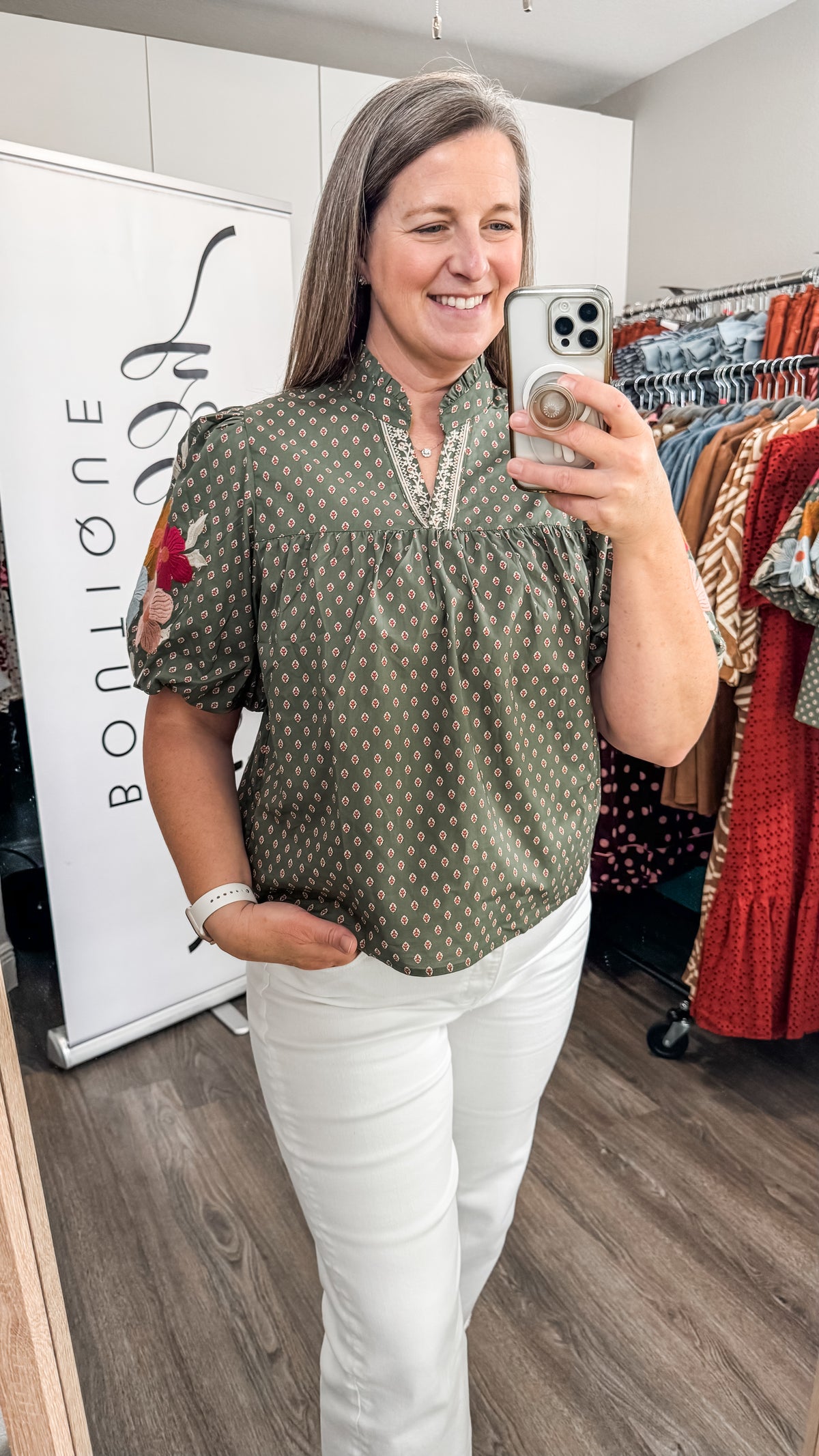 A woman takes a mirror selfie in a boutique while wearing a short-sleeve olive green blouse with a small white geometric pattern and white pants. A clothing rack with colorful garments and a white sign reading 'Bon Ligne' are visible in the background.