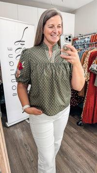 A woman takes a mirror selfie in a boutique while wearing a short-sleeve olive green blouse with a small white geometric pattern and white pants. A clothing rack with colorful garments and a white sign reading 'Bon Ligne' are visible in the background.
