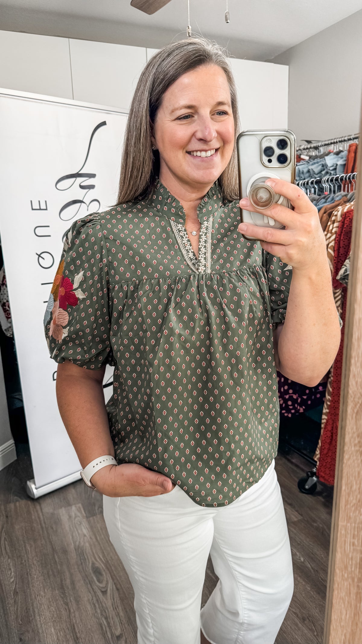 A woman takes a mirror selfie in a boutique, wearing a short-sleeve olive green blouse with a white geometric pattern, white pants, and holding a silver smartphone.