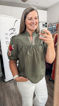 A woman takes a mirror selfie in a boutique, wearing a short-sleeve olive green blouse with a white geometric pattern, white pants, and holding a silver smartphone.