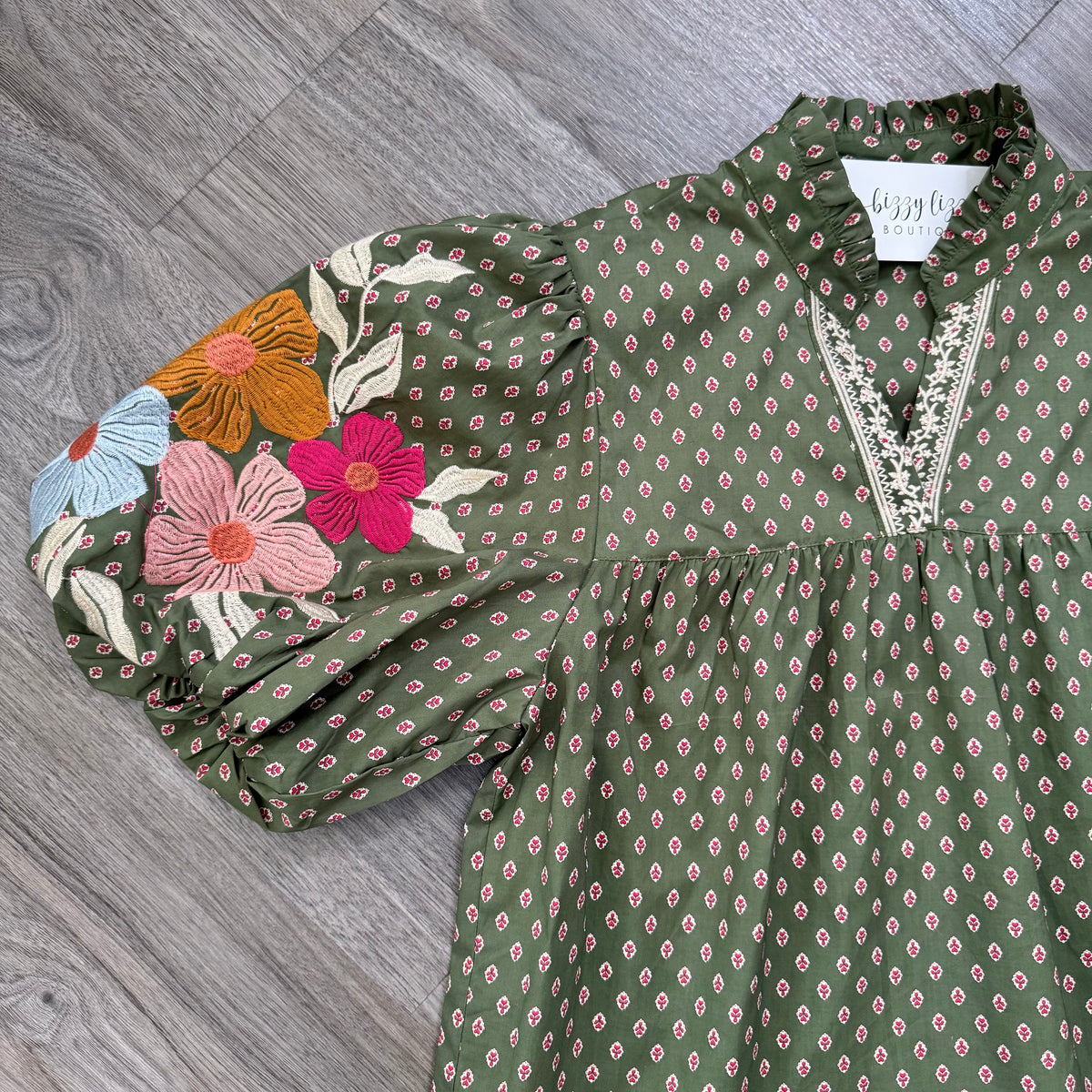 An olive green short-sleeve blouse with a small pink and white geometric print, featuring a lace-trimmed V-neckline and a colorful floral embroidery on the right sleeve, laid flat on a gray wood floor.
