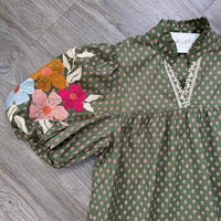An olive green short-sleeve blouse with a small pink and white geometric print, featuring a lace-trimmed V-neckline and a colorful floral embroidery on the right sleeve, laid flat on a gray wood floor.