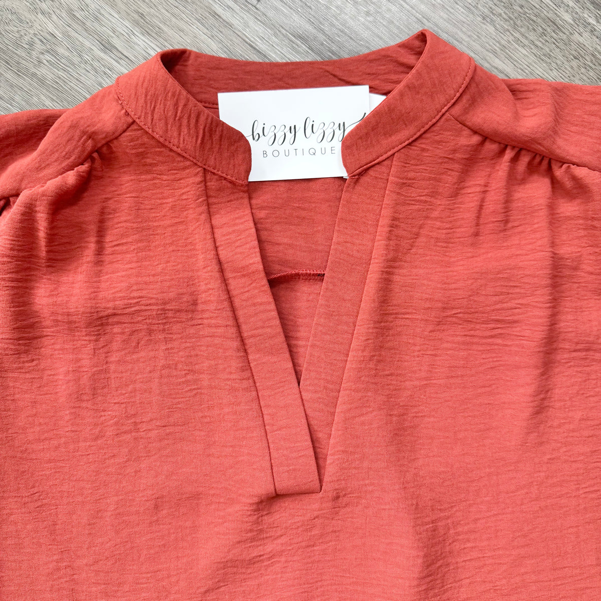 A close-up of a coral pink crinkled blouse with a split V-neckline and shoulder puff details, featuring a white neck label that reads 'bizzy lizzy BOUTIQUE'.