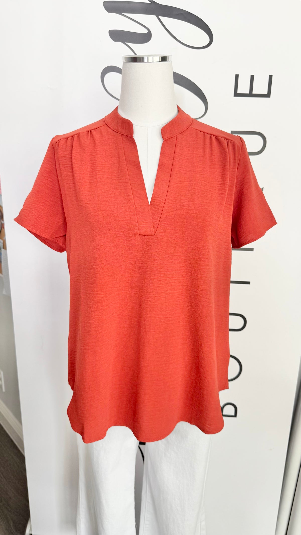A short-sleeve coral orange blouse with a split V-neckline burnt orange top, displayed on a white mannequin torso against a white boutique backdrop.