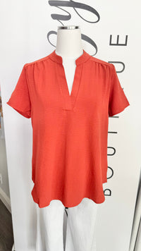 A short-sleeve coral orange blouse with a split V-neckline burnt orange top, displayed on a white mannequin torso against a white boutique backdrop.