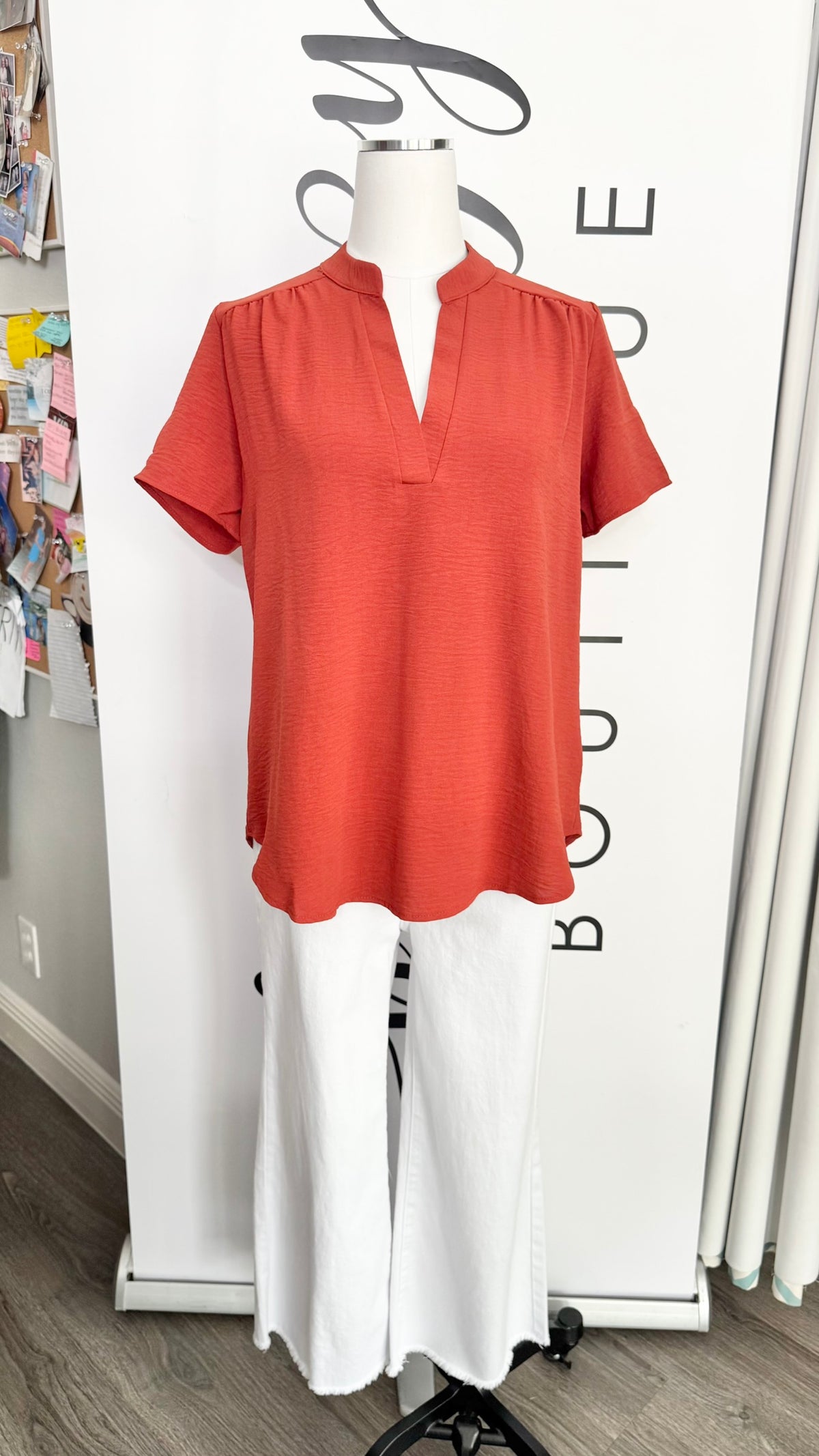 A terracotta orange short-sleeve blouse with a split neckline, displayed on a white mannequin torso paired with white wide-leg pants in front of a boutique backdrop.