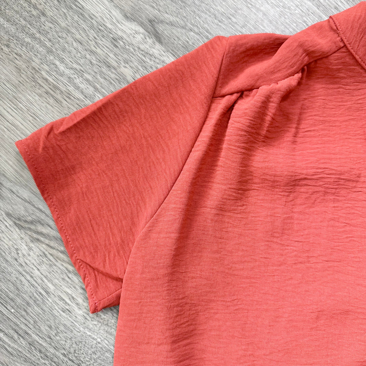 A close-up of a coral pink short-sleeve top with a textured crinkled fabric, laid flat on a gray wood-grain surface.