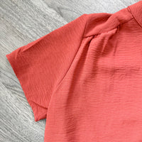 A close-up of a coral pink short-sleeve top with a textured crinkled fabric, laid flat on a gray wood-grain surface.