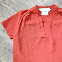 A short-sleeve crinkled cotton top in a muted terracotta or burnt orange color, laid flat on a gray wood-textured surface. The shirt features a split V-neckline with a small keyhole cutout at the center.