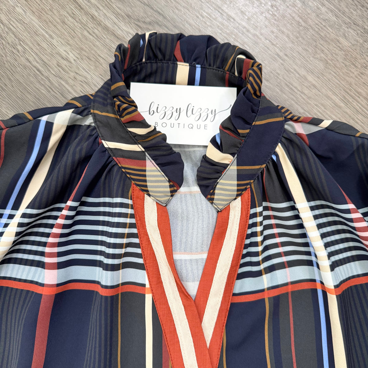 A close-up of a plaid and striped blouse from Bizzy Lizzy Boutique, featuring a navy blue base with red, beige, and light blue stripes, laid flat on a gray wood-textured surface.