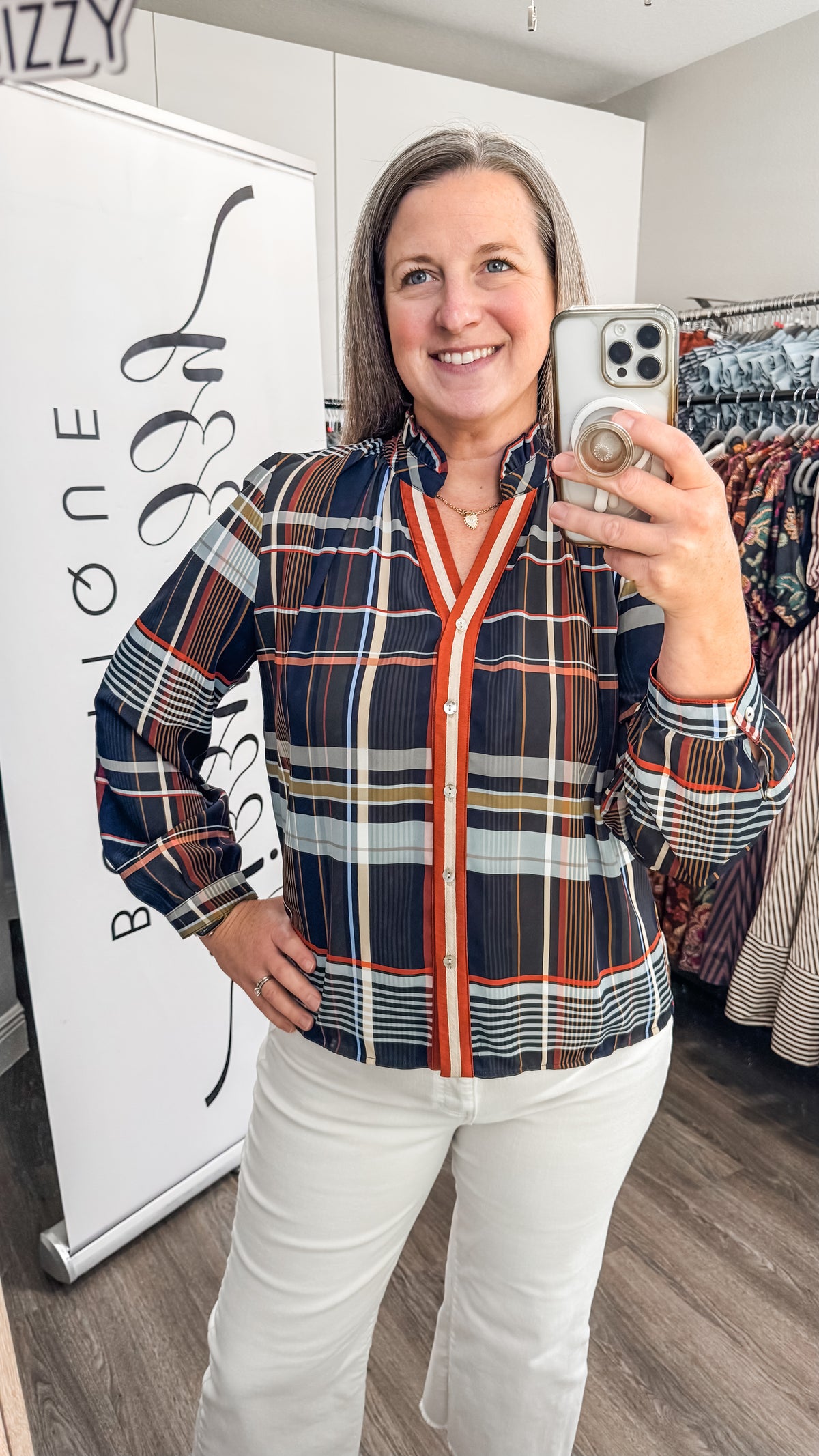 A woman takes a mirror selfie in a boutique while wearing a navy blue plaid button-up shirt with a bright orange-red center and white pants.