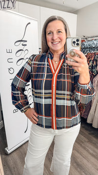 A woman takes a mirror selfie in a boutique while wearing a navy blue plaid button-up shirt with a bright orange-red center and white pants.