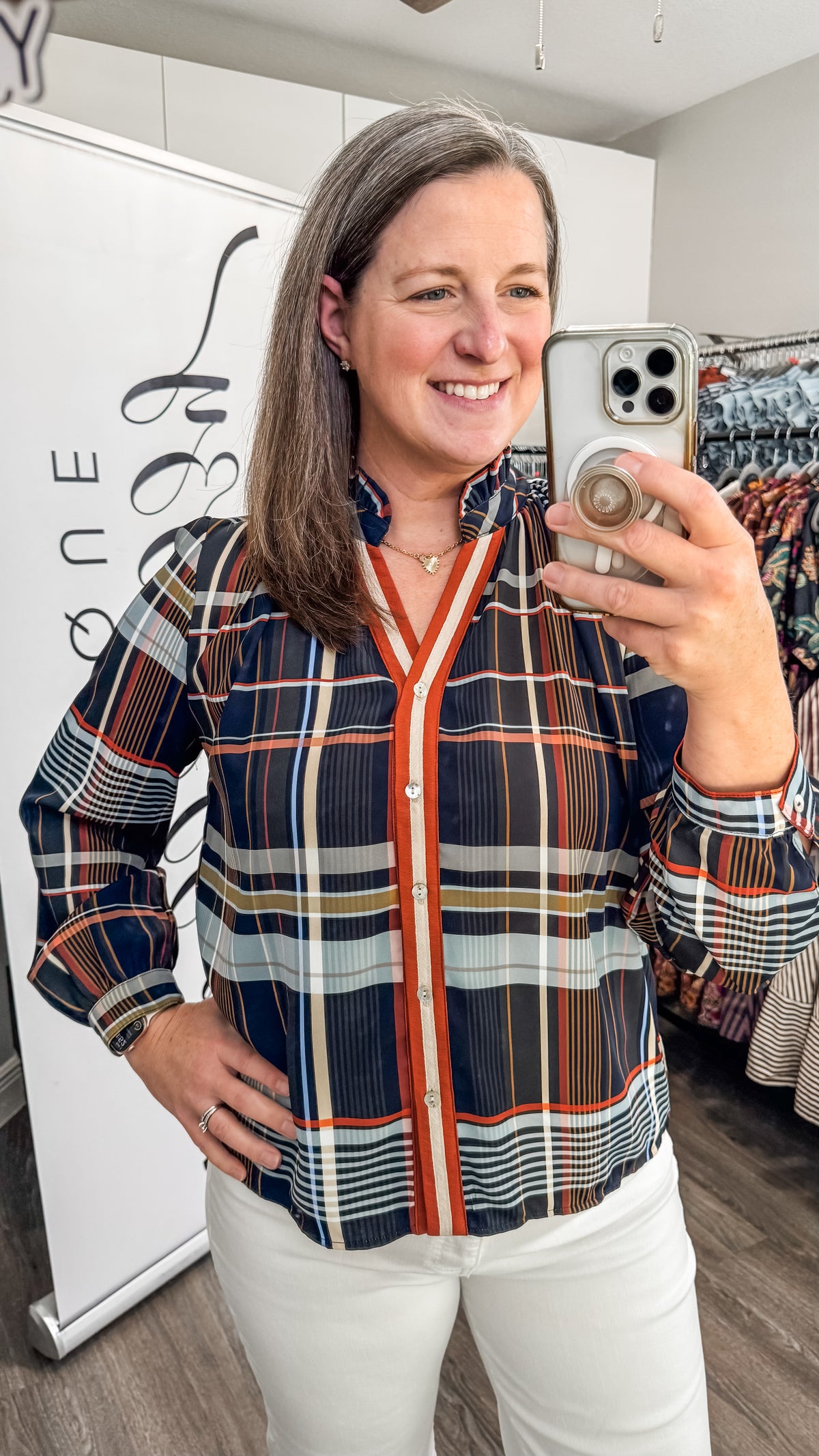 A woman takes a mirror selfie in a boutique while wearing a long-sleeve plaid button-up blouse with navy, orange, white, and gray stripes, paired with white pants.