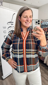A woman takes a mirror selfie in a boutique while wearing a long-sleeve plaid button-up blouse with navy, orange, white, and gray stripes, paired with white pants.