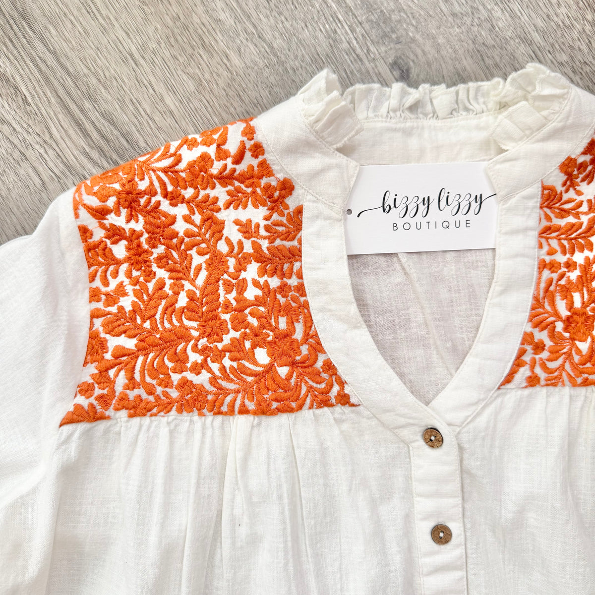 White blouse with orange floral embroidery on a wooden surface, featuring 'bizzy lizzy boutique' label.