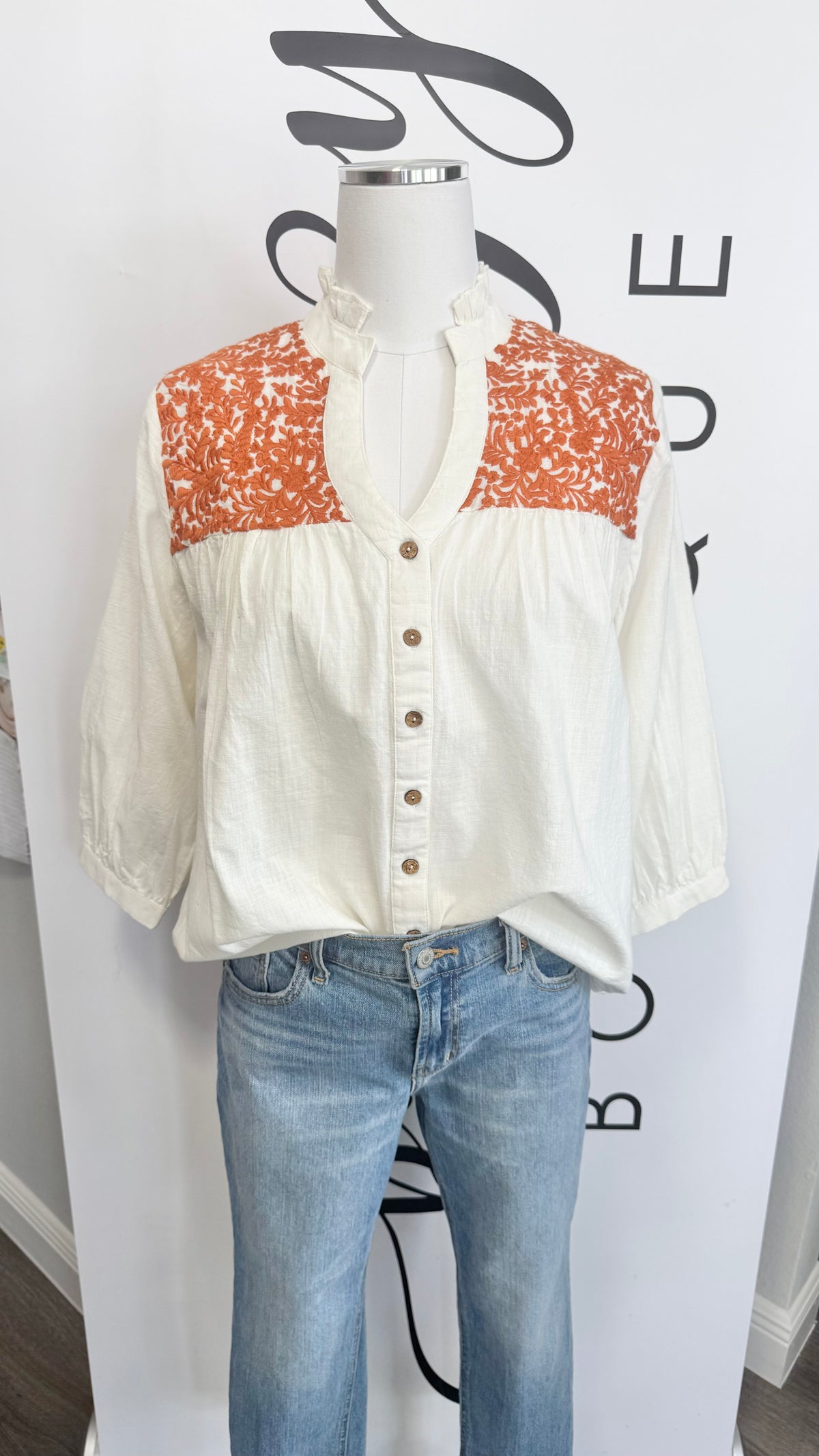 Mannequin wearing a white blouse with orange floral accents and blue jeans against a white background.