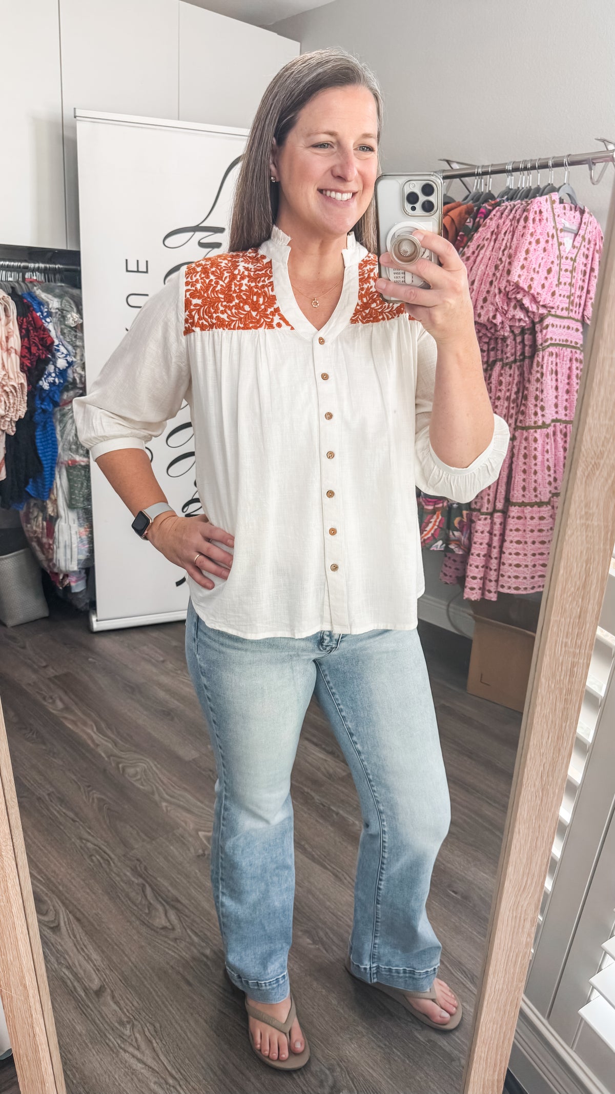 Woman taking a mirror selfie wearing a white blouse with orange accents and blue jeans in a clothing store.