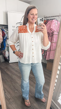 Woman taking a mirror selfie wearing a white blouse with orange accents and blue jeans in a clothing store.
