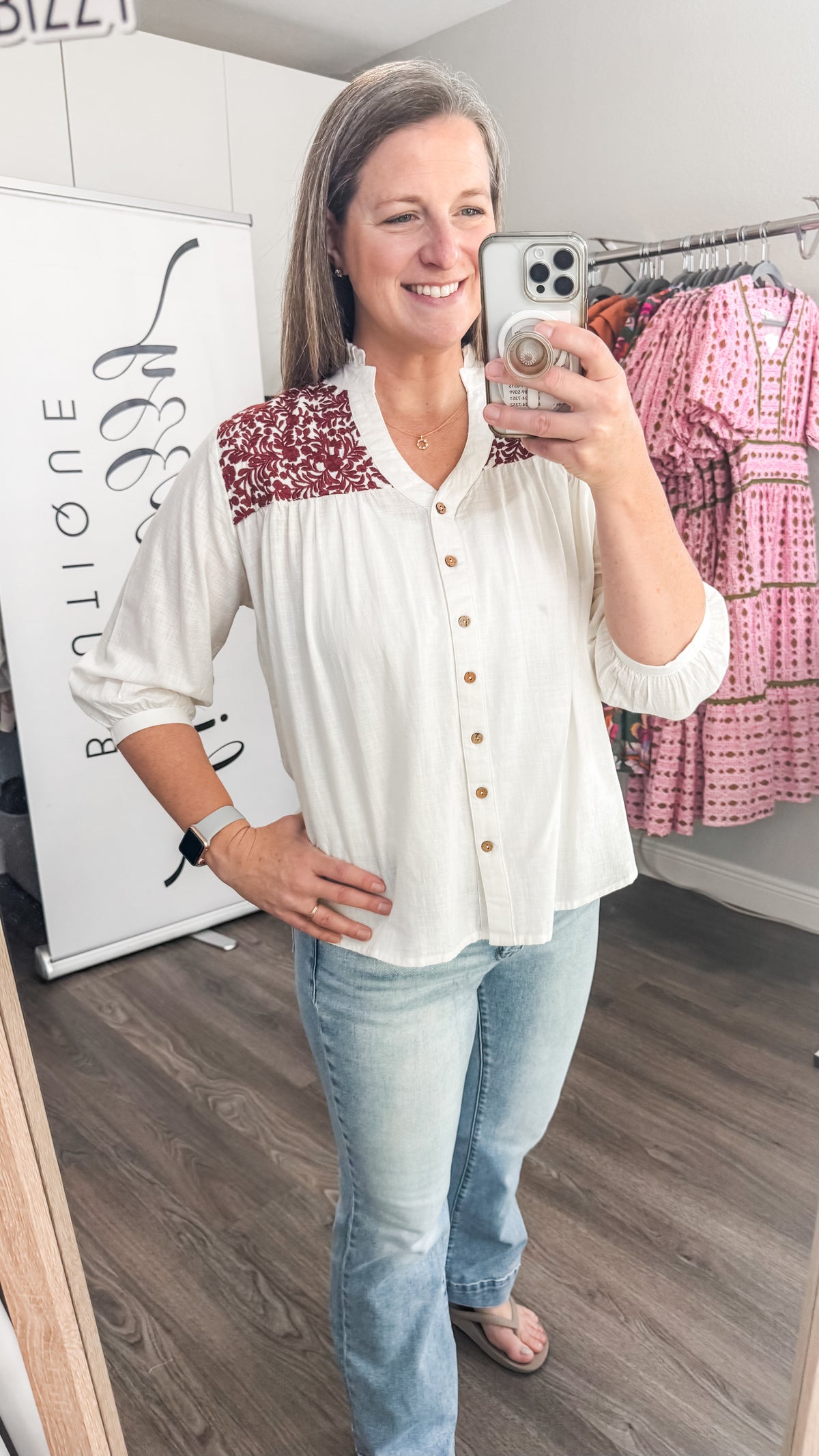 Woman taking a mirror selfie wearing a white blouse with Maroon embroidery and light blue jeans in a store.