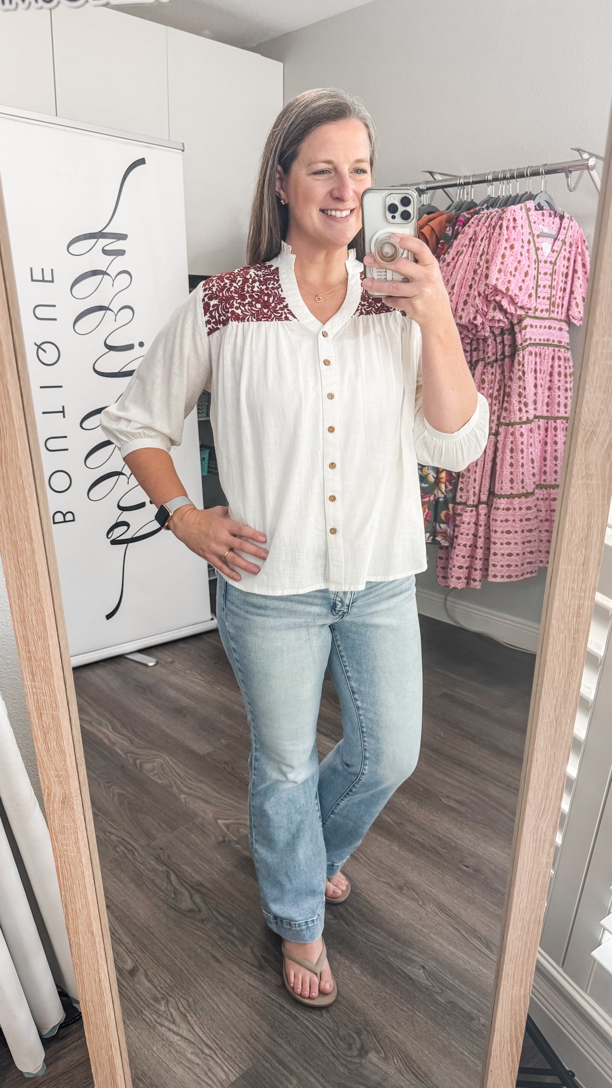 Woman taking a mirror selfie wearing a white and maroon embroidered blouse and light blue jeans in a room with clothing racks.