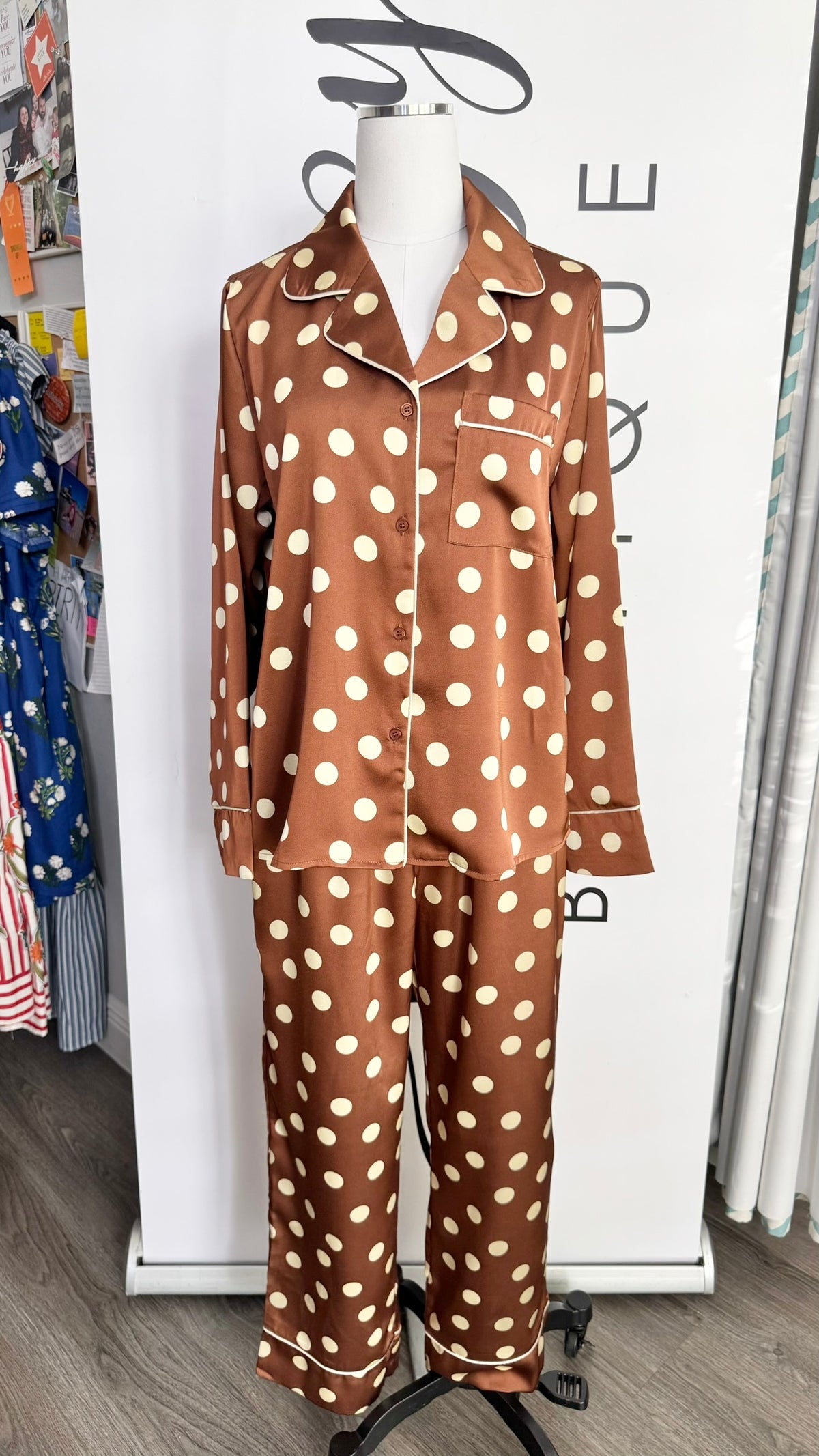 A two-piece pajama set displayed on a mannequin torso in a retail setting. The pajama set consists of a long-sleeve button-up top and matching pants in a brown satin-like fabric with a cream polka dot pattern.