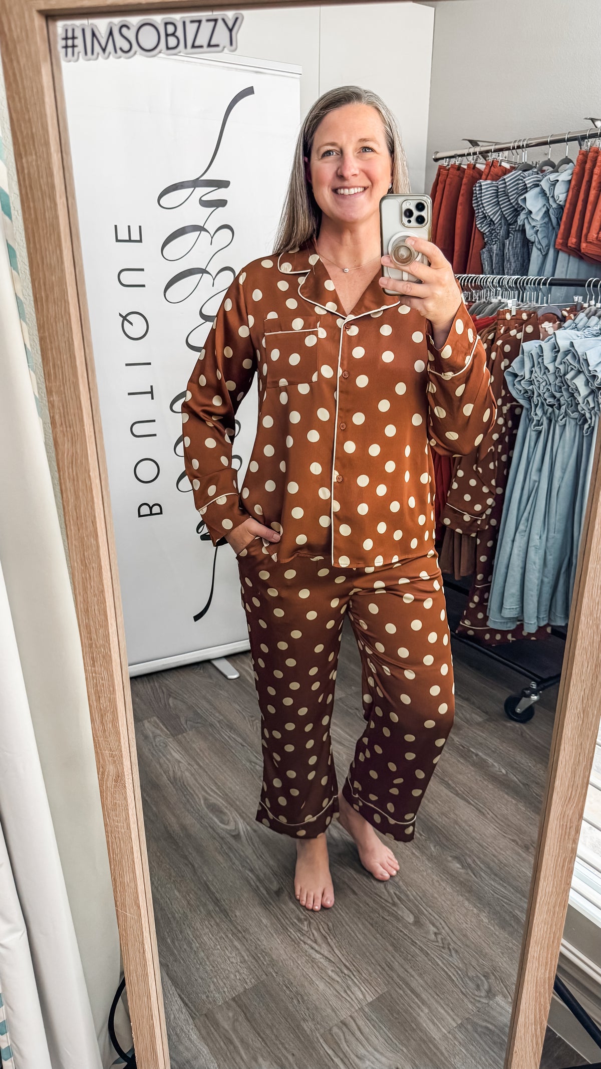 A woman takes a mirror selfie in a boutique wearing a rust-brown and white polka-dot pajama set. 