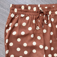 Close-up of brown pants with a white polka dot pattern and an elastic drawstring waistband, laid flat on a gray wood-textured surface.