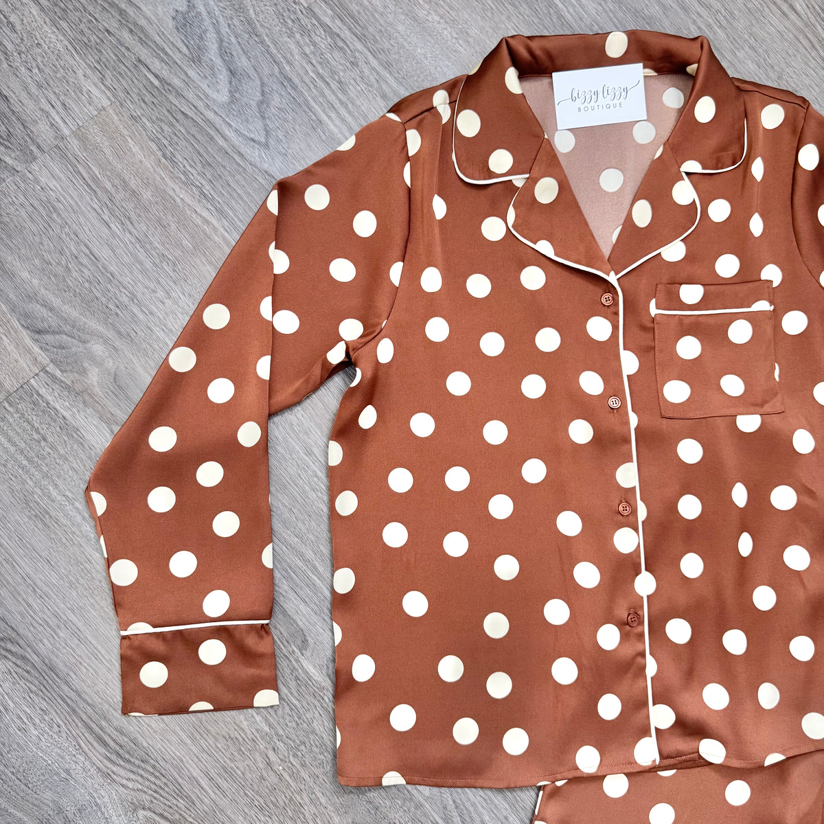 A long-sleeve pajama shirt in brown with white polka dots and white piping, laid flat on a gray wood-textured surface.