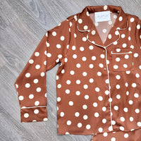 A long-sleeve pajama shirt in brown with white polka dots and white piping, laid flat on a gray wood-textured surface.
