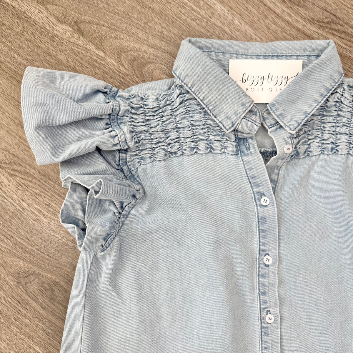 Light blue denim shirt with ruffled sleeves on a wooden surface, featuring a Bizzy Lizzy Boutique brand tag.