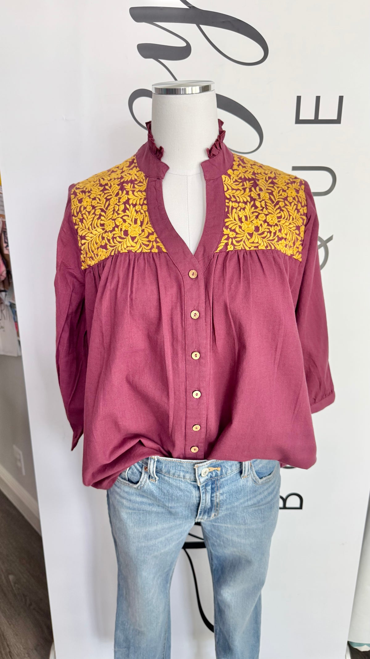 Mannequin wearing a maroon blouse with yellow embroidery details and blue jeans against a white background.