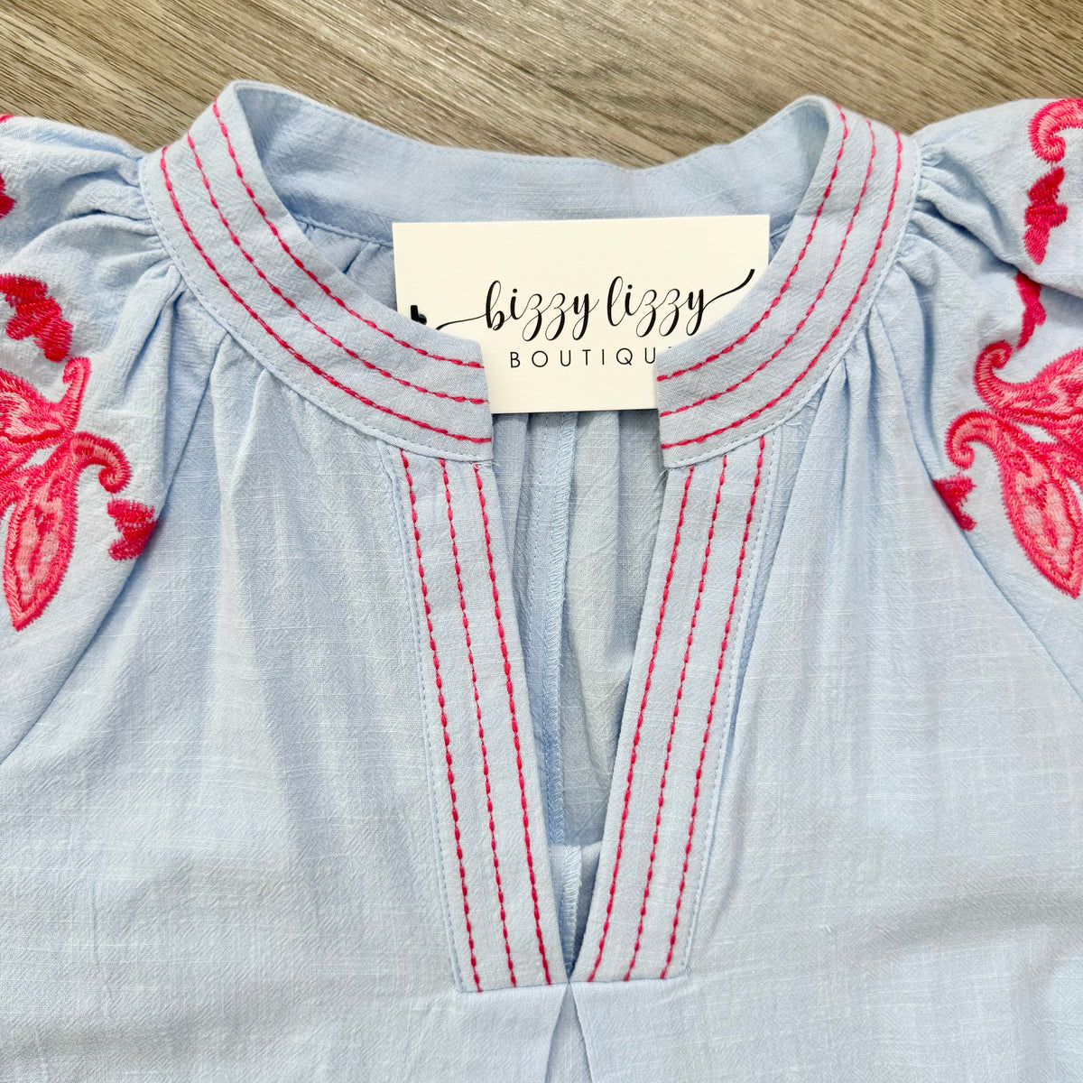 Light blue blouse with red embroidery and 'bizzy lizzy boutique' label on a wooden surface