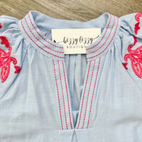 Light blue blouse with red embroidery and 'bizzy lizzy boutique' label on a wooden surface