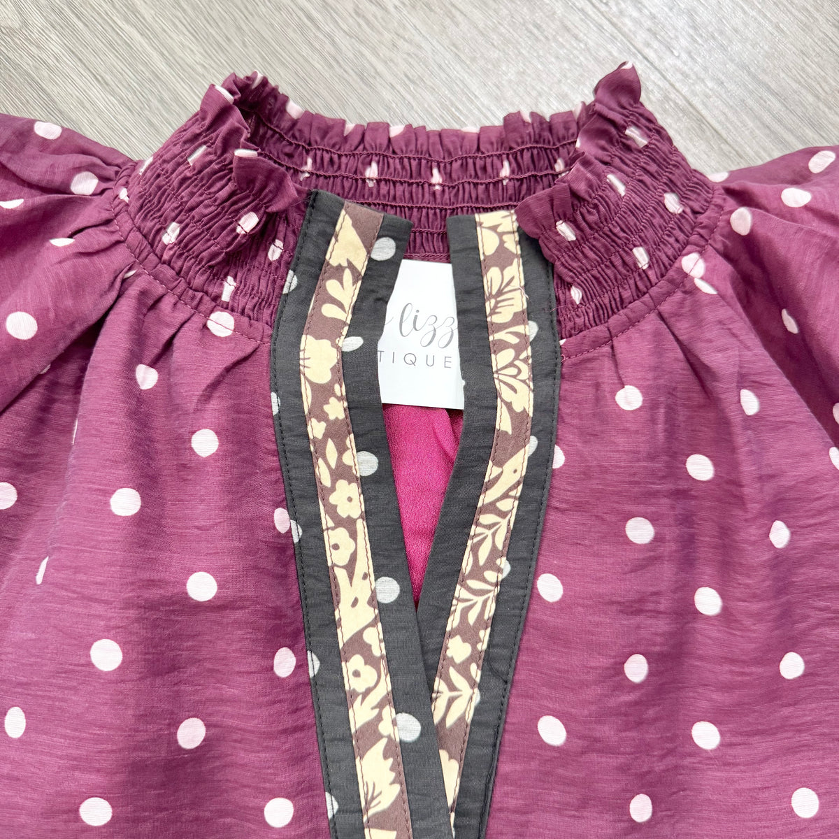 Close-up of a dusty maroon smocked blouse with white polka dots, featuring a decorative split neckline with dark gray, beige, and brown floral patterned trim. 