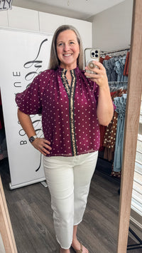 A woman takes a mirror selfie in a boutique wearing a burgundy short-sleeve blouse with white polka dots, paired with white pants.