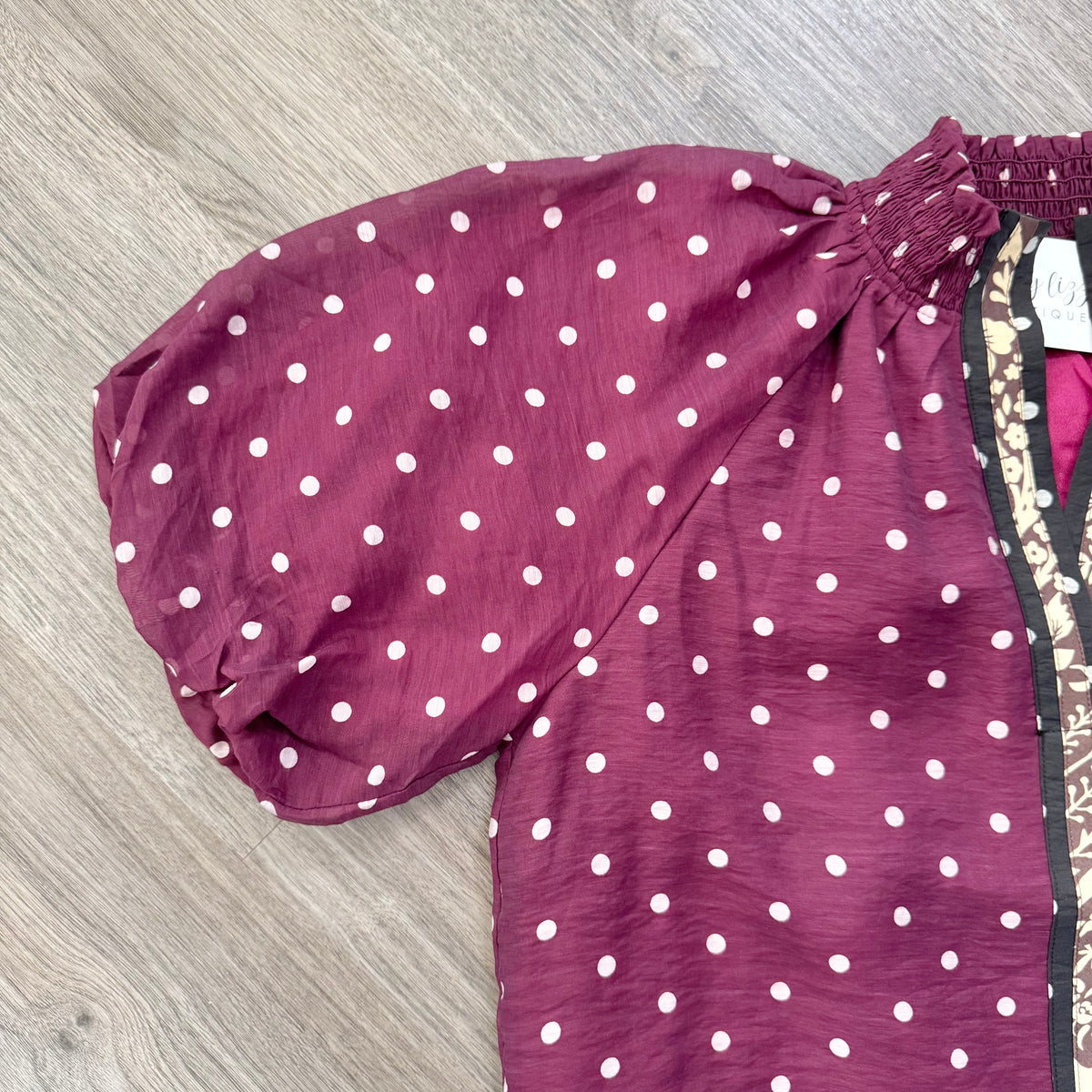 A close-up of a burgundy top with white polka dots, featuring a smocked elasticated yoke and a patterned side panel, laid flat on a gray wood floor.